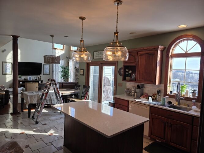 Bright kitchen with wooden cabinets, an island, and two pendant lights; adjacent dining and living areas with a ladder and covered furniture visible. Large window lets in natural light.