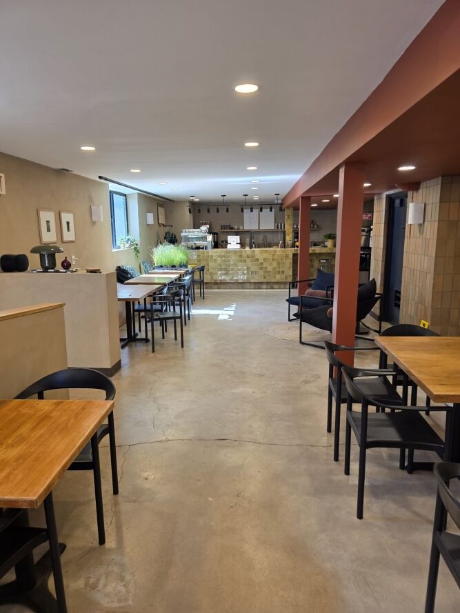 A modern cafe interior with wooden tables, black chairs, a concrete floor, and a view of the counter and seating area in the background.