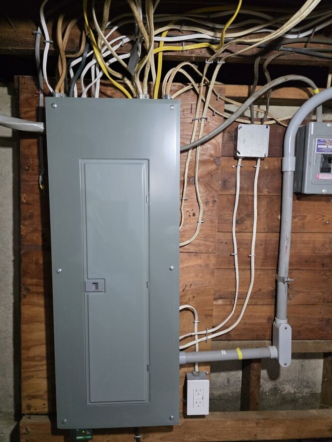 A gray electrical panel with multiple wires is mounted on an unfinished wooden wall, next to a utility box and an electrical outlet.