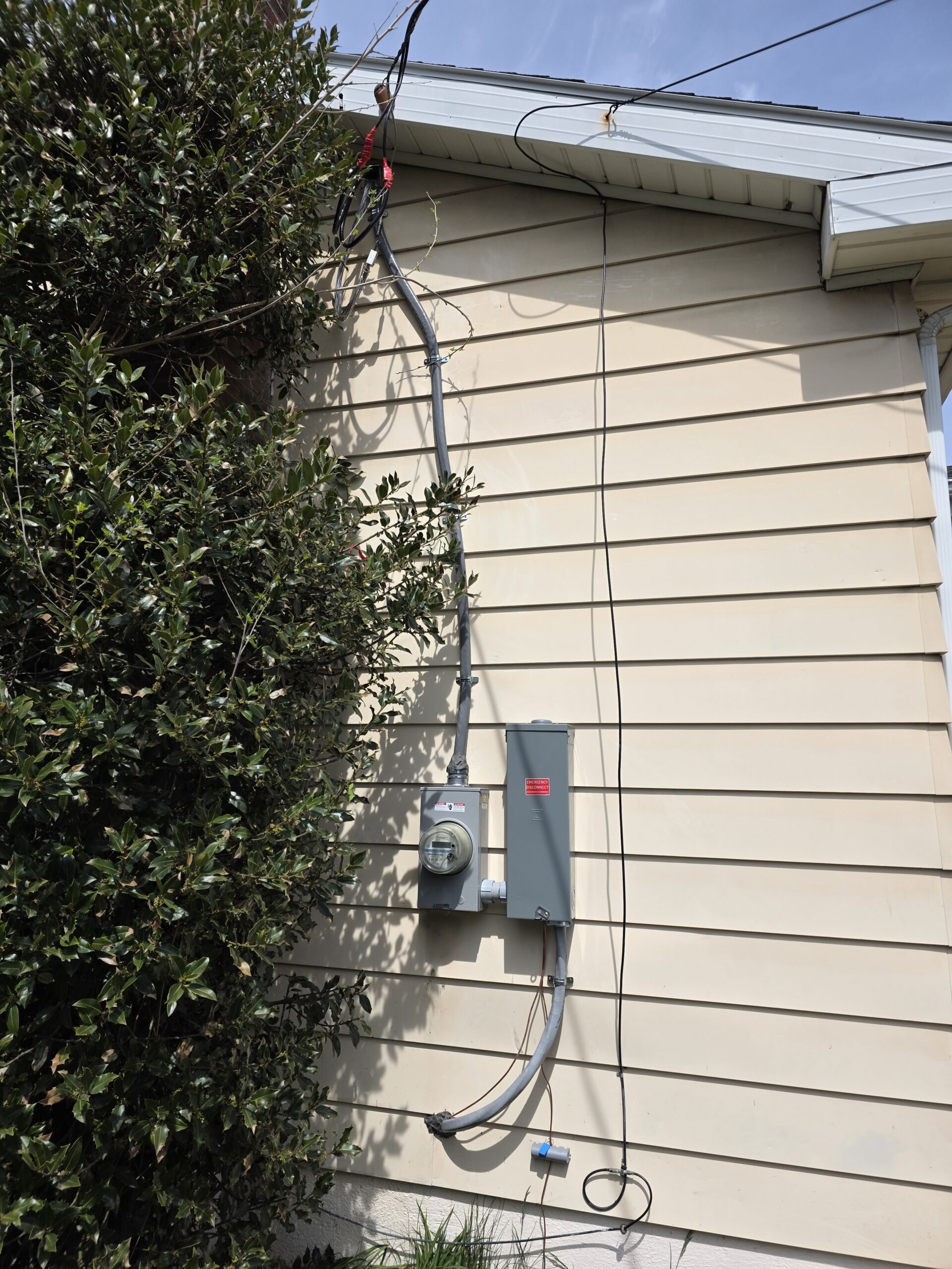Electric meter and breaker box mounted on the exterior wall of a house with overhead power lines and nearby bushes.