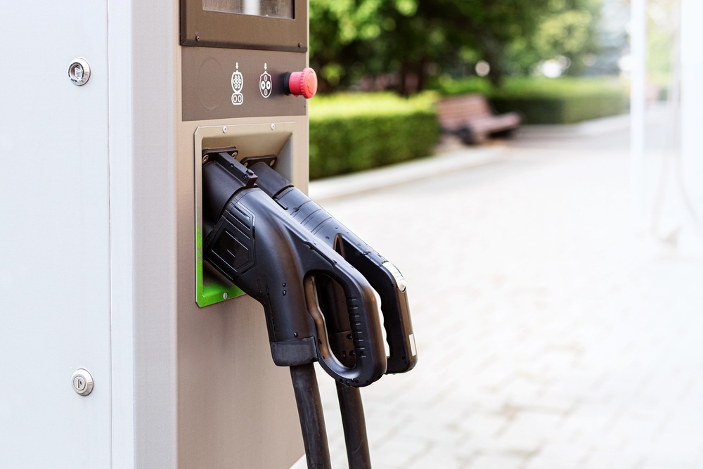 Close-up of an electric vehicle charging station with two charging plugs inserted, located outdoors near a paved walkway and greenery.