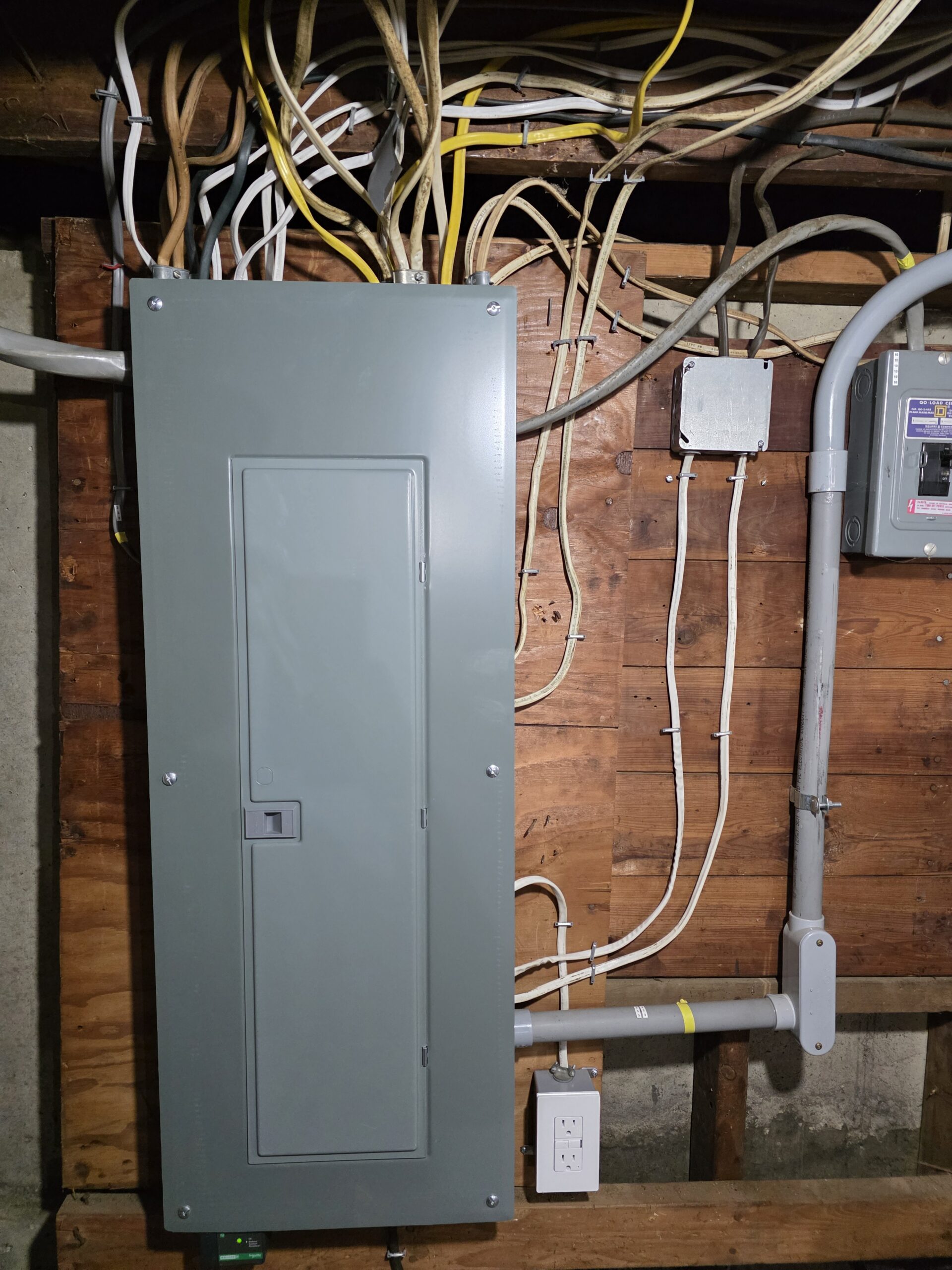 A gray electrical panel with multiple wires is mounted on an unfinished wooden wall, next to a utility box and an electrical outlet.