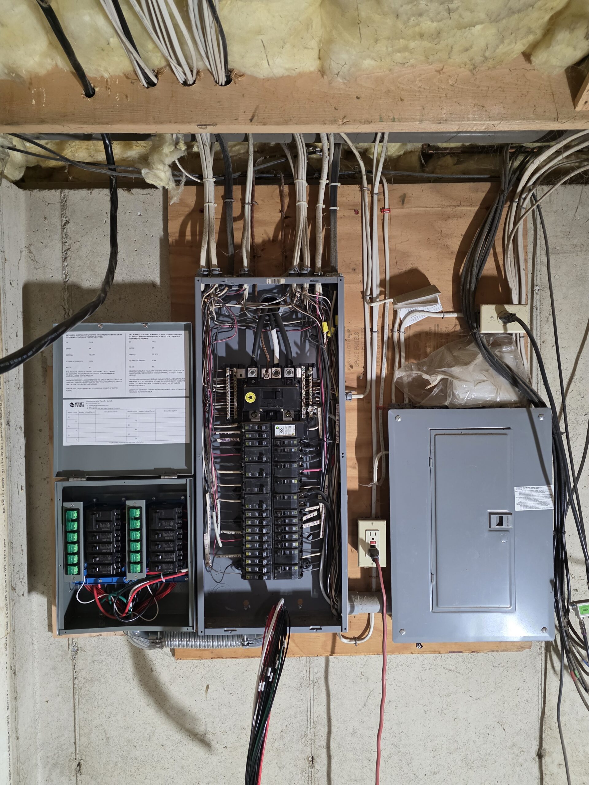 An open electrical panel with exposed wiring and circuit breakers mounted on a basement wall, next to a closed gray electrical box.