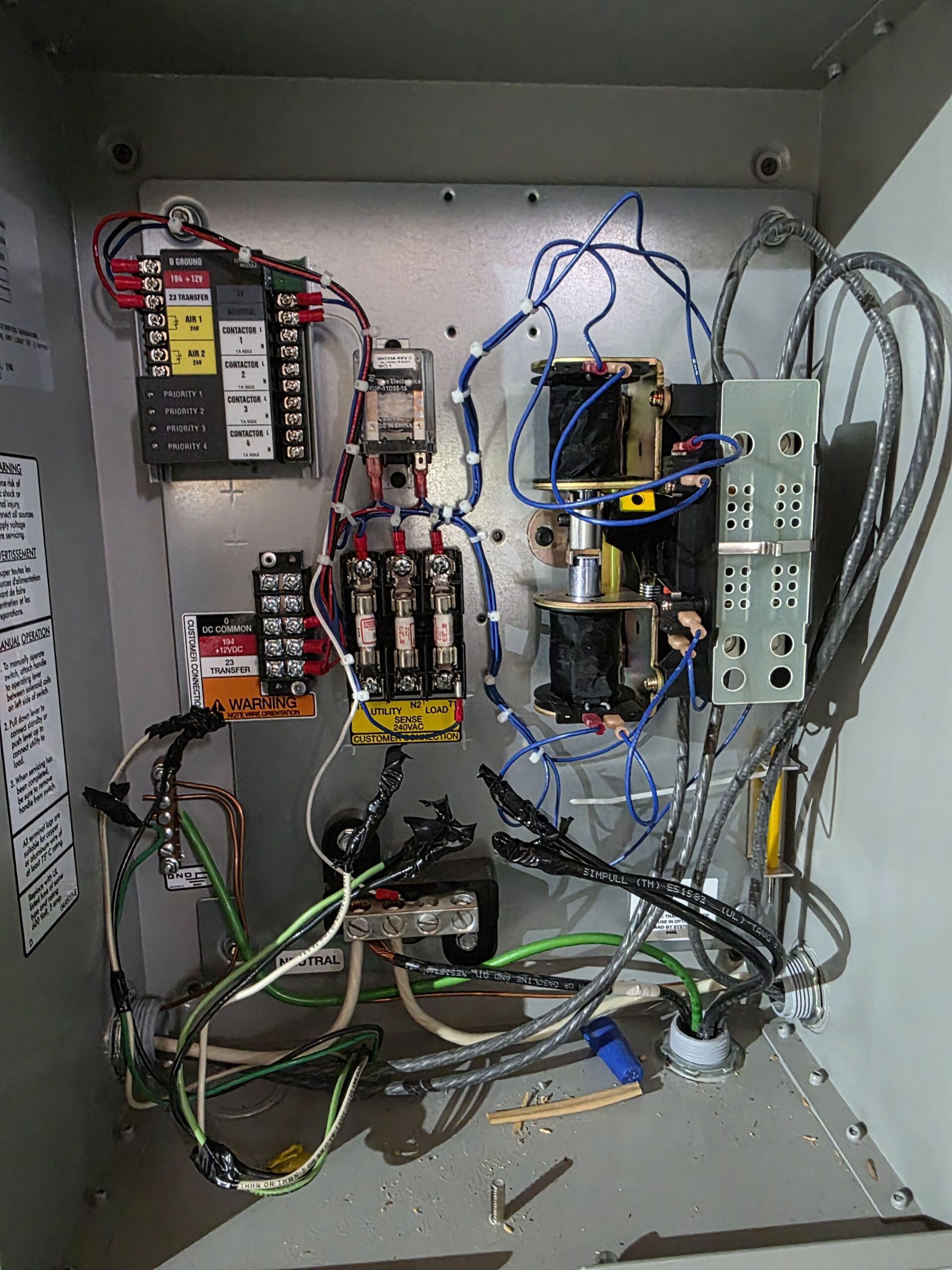 An open electrical control panel showing various wires, relays, switches, terminal blocks, and warning labels inside a metal enclosure.