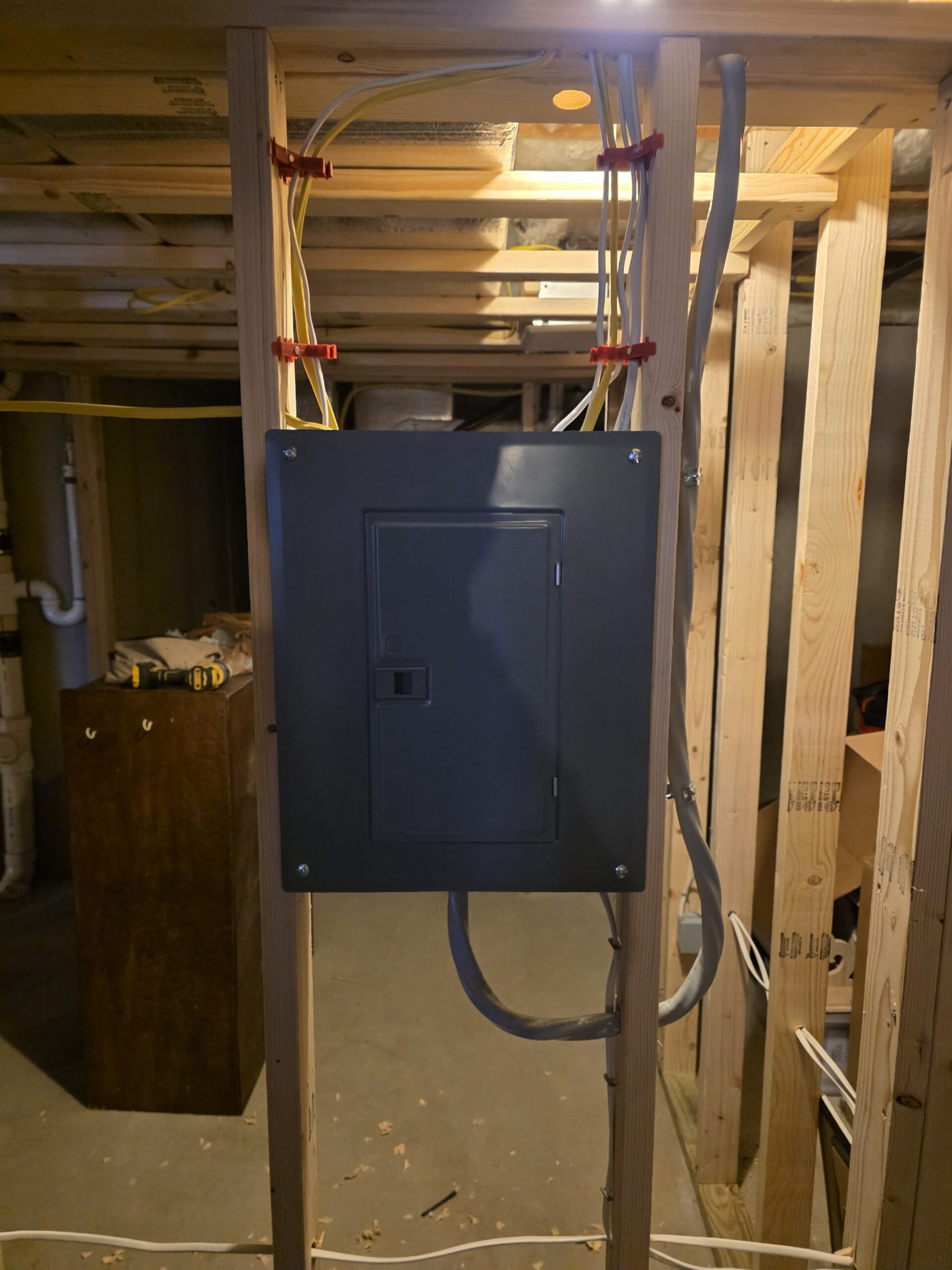 A gray electrical panel is mounted on wooden framing in a basement, with multiple wires entering from the top and bottom.
